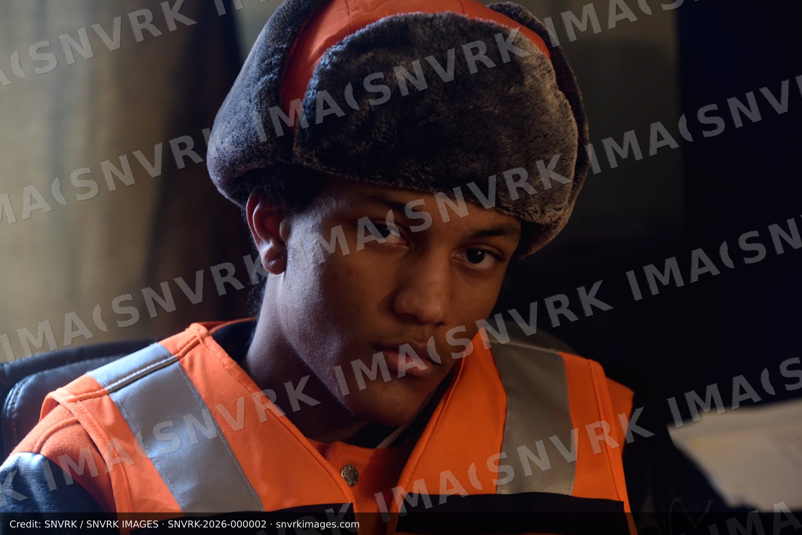 Young man in a faux-fur trapper hat and reflective orange vest over a leather jacket looks at the camera in warm low light.
