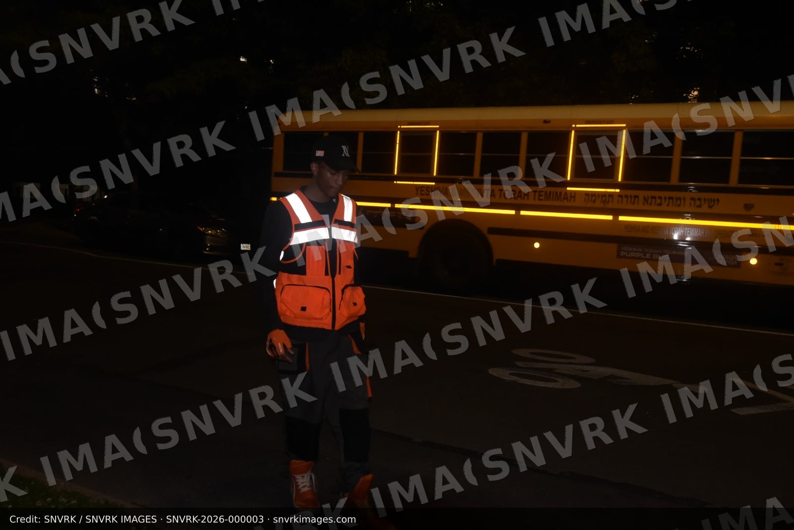 Young man in an orange reflective hi-vis vest, black cap, work gloves and orange high-top sneakers walks along a dark street at night in front of a parked yellow school bus. (file photograph)