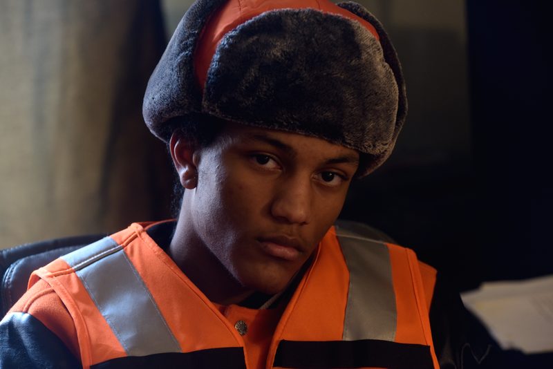 Young man in a faux-fur trapper hat and reflective orange vest over a leather jacket looks at the camera in warm low light.