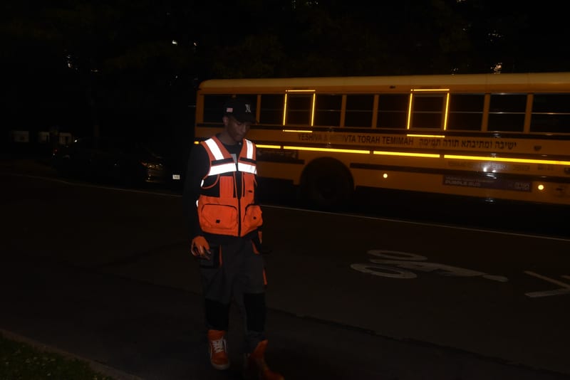 Young man in an orange reflective hi-vis vest, black cap, work gloves and orange high-top sneakers walks along a dark street at night in front of a parked yellow school bus.