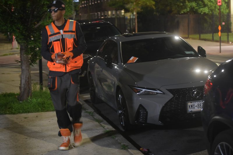 Man in an orange hi-vis vest, work gloves and high-top sneakers walks past a ticketed grey Lexus on a city sidewalk at night.